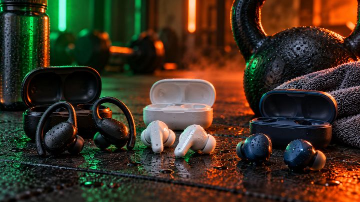 Three pairs of wireless sports earbuds with charging cases on a wet gym floor next to a kettlebell, a towel and a water bottle
